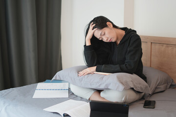 Fototapeta premium A young asian teenager struggle doing homework with tablet and notebook white sitting on bed, learning at home and getting headache. people struggle with technology and try hard to pass the exam.