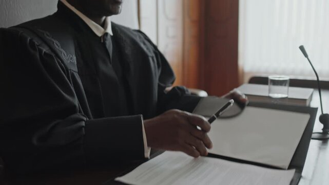 Tilt up shot of Black male judge hitting sound block with wooden gavel, opening folder and announcing start of court trial