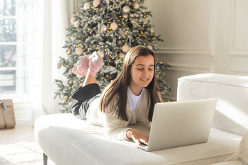 Smiling attractive young girl relaxing in front of a decorated Christmas tree reclining on a comfortable sofa working on her laptop computer