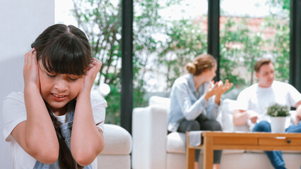 Stressed and unhappy young girl huddle in corner, cover her ears blocking sound of her parent arguing in background. Domestic violence at home and traumatic childhood develop to depression. Synchronos