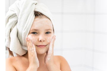 Portrait of a cute little girl in her pre-teens in the mirror, with cleansing foam on her face. Prevention of acne and first pimples on the skin.