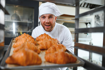 Handsome baker in uniform holding tray full of freshly baked croissants at the manufacturing