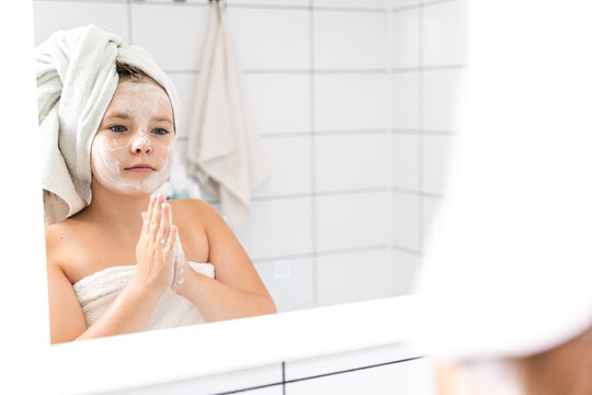 Portrait of a cute little girl in her pre-teens in the mirror, with cleansing foam on her face. Prevention of acne and first pimples on the skin.