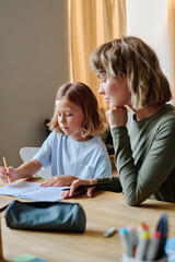 Fototapeta premium Young blonde mother sitting next to her lovely daughter while they doing homework