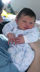 Newborn baby in a light blue onesie, being held by an adult, displaying a curious and alert expression, tender moment of early life and parental care