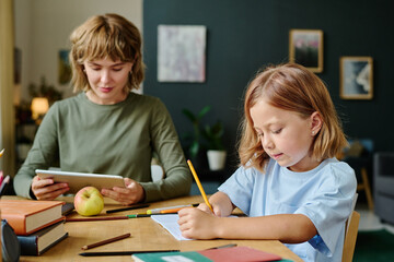 Little girl sitting at wooden table and drawing with yellow pen while her young mother reading...