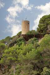 A tall historic tower stands on a hill with trees in the background. The sky is blue and there are clouds. Castle Sant Joann, Lloret de Mar, Spain. Vertical