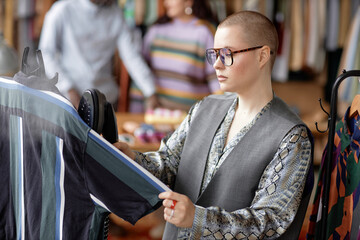 Medium side view shot of fashionable female shop assistant with buzz cut dressed in vintage clothes steaming second hand shirt at thrift store