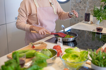 Elderly woman at home cooking vegetables in a pan while adding some pepper. Elderly lady in the kitchen preparing dinner. Healthy eating concept