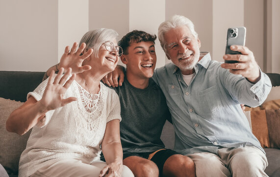 Happy multigenerational family of white-haired grandparents and young grandson on video call via mobile phone sitting together at home. Old and new generation waving