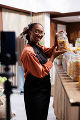 Selective focus on African American female vendor holding glass jar with pasta, filming social media content for the store. Black woman with an apron making a vlog to promote fresh, organic products.