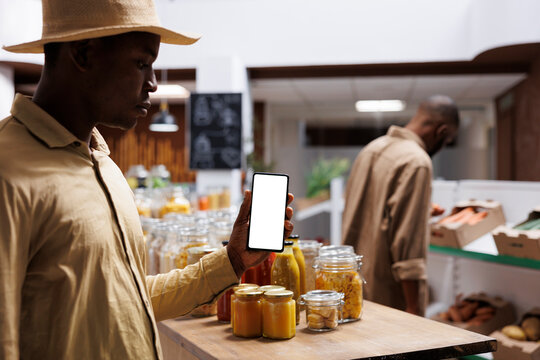 At an eco friendly grocery store, an African American male customer clutches a cell phone with an isolated white screen. The image shows a black man holding a mobile device with a chromakey template.