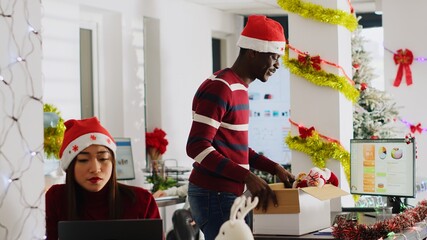 Exicted african american employee clearing desk before leaving for vacation, dancing around festive decorated office. Man wearing Santa hat taking days off to go on xmas holiday, feeling merry