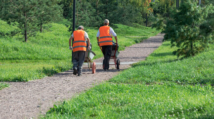 Fototapeta premium Two men in orange vests are pushing wheelbarrows down a path