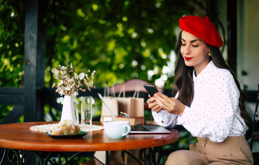 Gorgeous smiling cheerful brunette woman in a red beret resting and having breakfast in city cafe sitting at table on the terrace and using her smart phone for shopping online or chatting