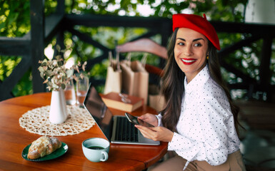 Gorgeous smiling cheerful brunette woman in a red beret resting and having breakfast in city cafe sitting at table on the terrace and using her smart phone for shopping online or chatting