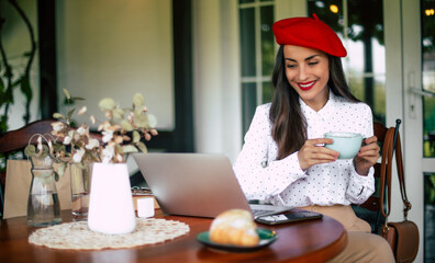 Stylish beautiful smiling cheerful brunette woman in a red beret resting and having croissant breakfast with coffee in city cafe sitting at table on the terrace with phone and laptop on the table