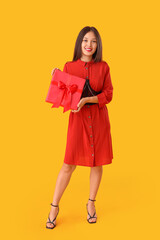 Beautiful young Asian woman with gift box on yellow background