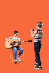 Little boy recording his brother playing guitar on orange background