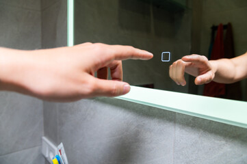 Woman's hand turns on the illumination of the bathroom mirror by touching the touch button on and...