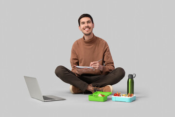 Young man with notebook and lunchbox sitting on grey background