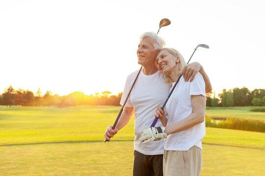 elderly senior couple in uniform holding golf clubs and standing on the background of golf course at sunset and smiling, old man and woman hugging and smiling while playing golf and looking at copy