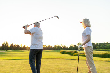 elderly senior couple in uniform playing golf on golf course at sunset, old man swinging club at...