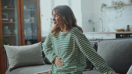 Young sick woman sitting on couch in living room clutching her stomach in pain, feeling burning sensation or cramps at home - Powered by Adobe