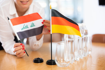 Little flag of Germany on table with bottles of water and flag of Iraq put next to it by positive young woman in meeting room