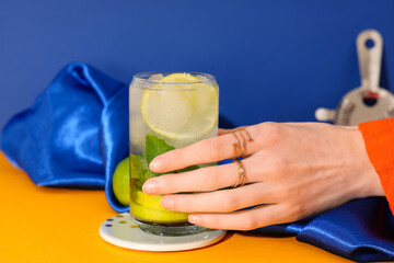 Woman taking glass of fresh mojito from orange table on blue background