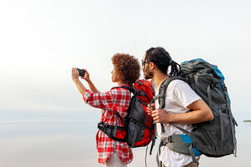 young african american couple with hiking backpacks and equipment standing against sea and sky and taking landscape photo on smartphone, curly woman and man tourists traveling and filming isolated