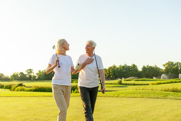 elderly senior couple in uniform holding golf clubs and walking on the background of golf course at sunset and talking, old man and woman walking and talking on golf game and looking at copy space