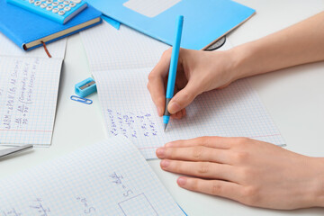 Woman writing math formulas in copybooks and stationery on light background, closeup