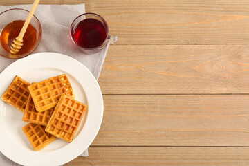 Plate with tasty Belgian waffles, bowl of sweet honey and glass cup of tea on wooden background