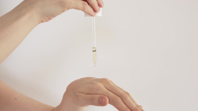 A hand using a dropper to apply a few drops of topical therapy serum to the back of the hand on a white background. A visual representation of skincare, medicine, and dermatology treatments