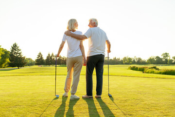 elderly senior couple in uniform playing golf on golf course at sunset, old man instructor teaching woman how to hold golf club and swing ball, grandfather helping grandmother play outdoors