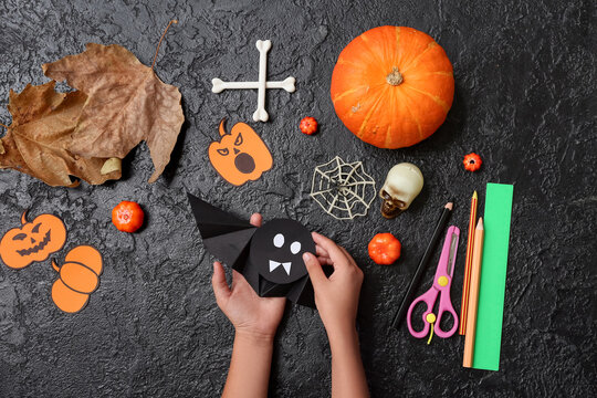Child with supplies making Halloween decorations and origami bat on grunge black background
