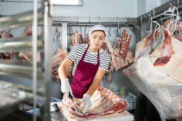 Female employee of local butcher shop divides large huge piece of meat carcass with knife