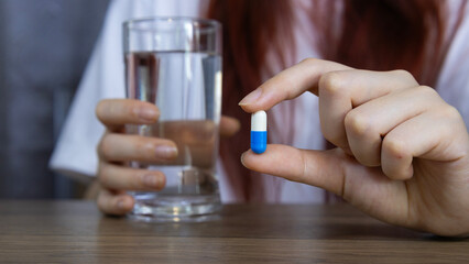 Asian woman's hand holding white-blue pill capsule carefully with fingers and holding glass of water, symbolizing healthcare, Concept of pharmacy, medicine, supplement, vitamin and medical treatment.