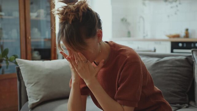Young miserable woman sitting on couch at home, feeling physical pain or suffering from depression