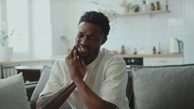 Young Black man sitting on a couch in the living room, holding his jaw with pained expression as having severe toothache at home