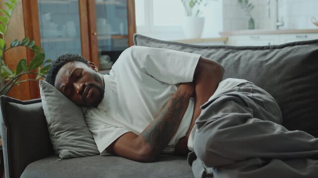 Young distressed African American man lying on sofa at home and holding stomach while feeling pain