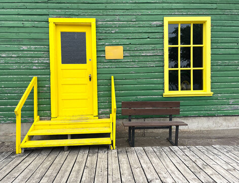 Fachada de casa con puerta y dos ventanas de madera en color amarillo brillante y piso de madera. 