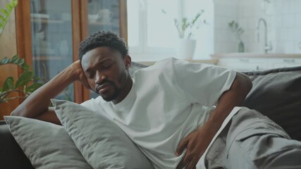 Young sick African American man with distressed expression on his face lying on sofa at home and experiencing stomach ache