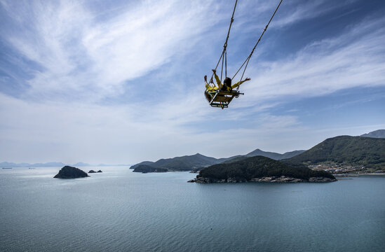 Namhae-gun, Gyeongsangnam-do, South Korea - May 14, 2022: A female is swinging with her arms spread in the air against sea and sky at Sulli Sky Walk