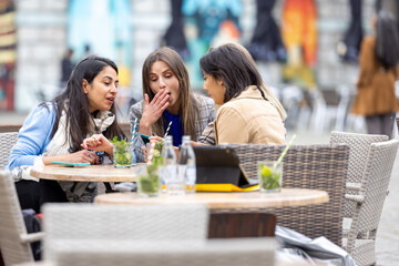 Three women are socializing over drinks at a cafe, using mobile devices and having a good time together outdoors