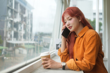 Fototapeta premium Attractive young woman with long red hair sitting by a large window, talking on mobile phone.