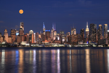 New York City skyline illuminated at dusk with orange full moon, view from Hudson River, USA