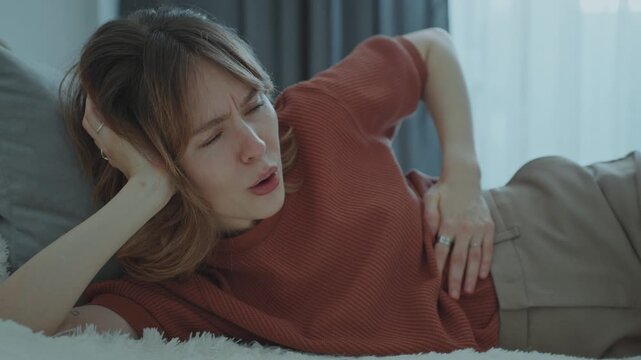 Young woman lying on bed at home, wincing and touching her stomach as feeling cramping and pain during periods
