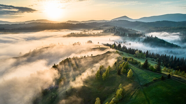 Aerial view of breathtaking sunrise over misty landscape with rolling hills and verdant fields. Soft light pierces through fog, casting long shadows and illuminating trees, creating magical scene.